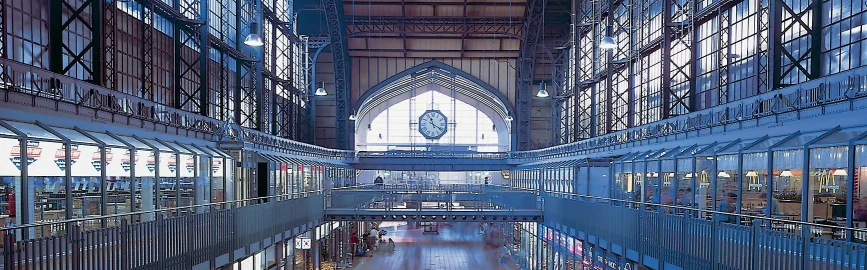 Eine zweistöckige Bahnhofshalle mit Blick vom Obergeschoss zum Eingang über dem eine analoge Uhr hängt.